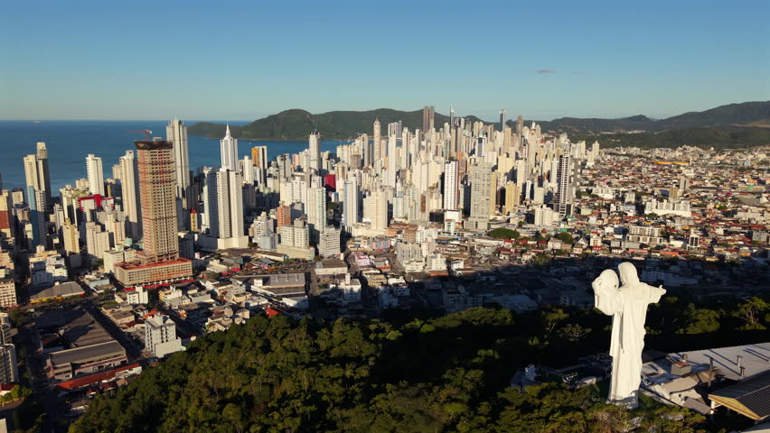 Smooth right-slider drone shot unveils Camboriú’s hilltop Christ statue, framed against morning-lit skyscrapers and ocean background. Ideal for spiritual or destination marketing.