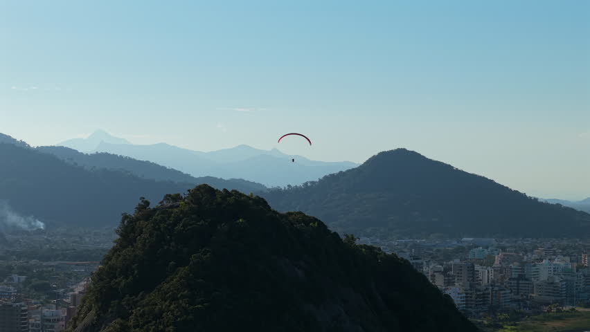 A stunning aerial parallax tracking shot follows a paraglider soaring peacefully over the beautiful mountains and city of Caiobá, Brazil, on a clear day.