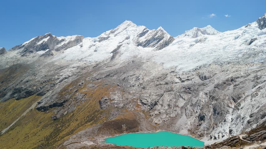 A majestic aerial shot reveals the stunning landscape of the Santa Cruz trek in Peru, showing a turquoise glacial lake at the base of snow-capped Andes mountains.