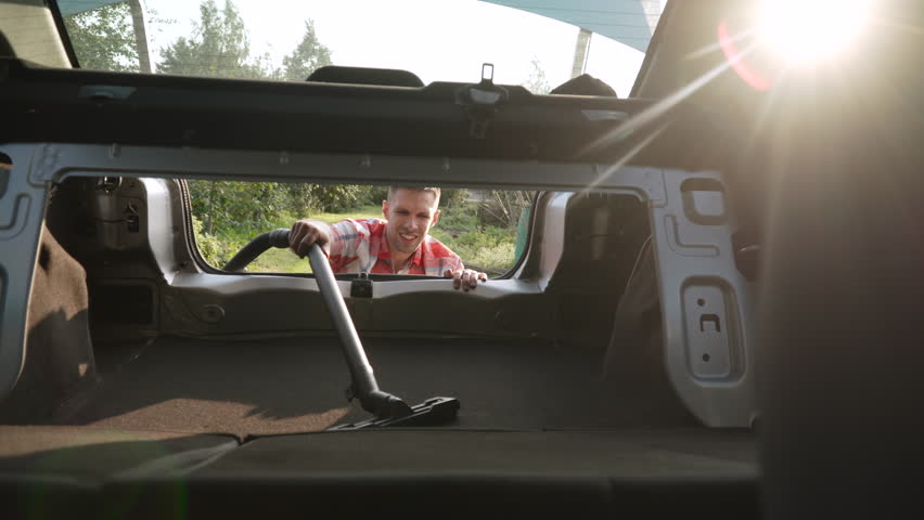 Smiling caucasian man vacuuming trunk of family car in sun rays. Portrait of happy car enthusiast cleaning interior of car on summer evening. Enjoyment from favorite job.