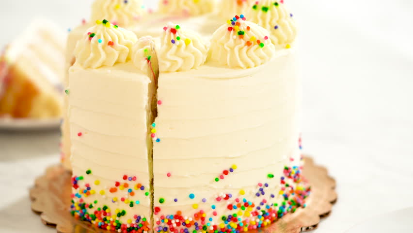 The birthday cake is positioned on a golden board with a slice missing, revealing fresh fruit filling between the layers. Bright sprinkles encircle the base of the frosting.
