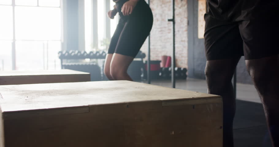 Diverse workout partners springing from low squat into plyo box jumps in gym, building leg strength. Fitness, training, athletic, movement, exercise, motivation, strength