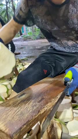 A worker peels a fresh green coconut using a sharp machete, showcasing a traditional tropical method to prepare refreshing coconut water and edible white flesh for culinary use.