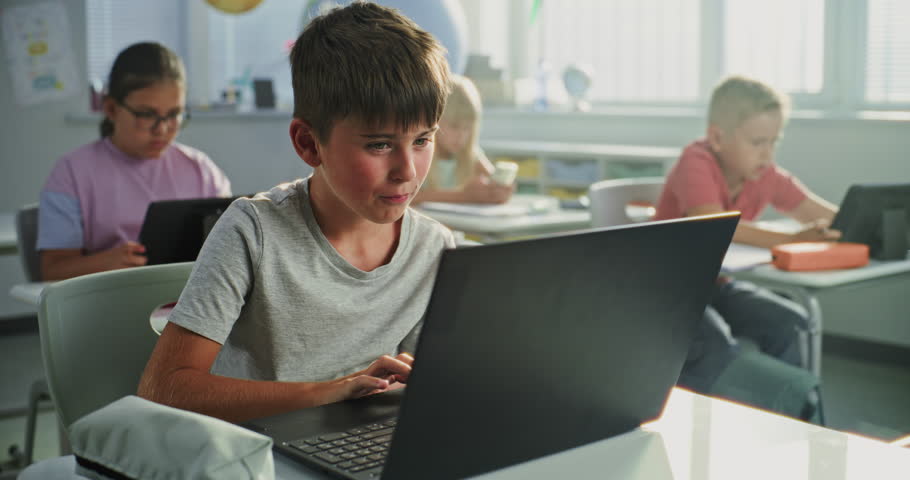 Elementary School Boy Sitting at Desk, Using Laptop in Classroom. Team of Smart Diverse Kids Working on Their Laptops and Tablet Computers, Studying Computer Science, Basic Programming and Coding.