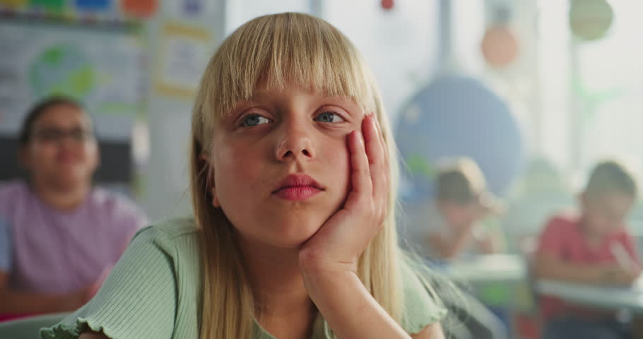 Close Up of Sad Primary School Girl Sitting at Desk, Getting Tired During Lesson or Upset Because of Bad Grade on Homework. Group of Young Children Learning Science or Geography in Modern Classroom.