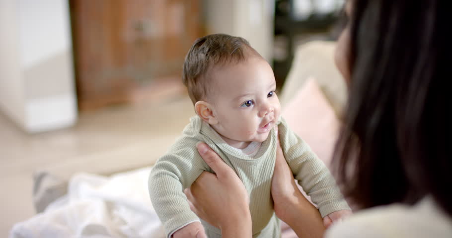 Mother noticing daughter gaze and lifting her to sit on blanket, guiding head turns in bedroom. Caregiver, bonding, tenderness, nurturing, affection, domestic, cozy
