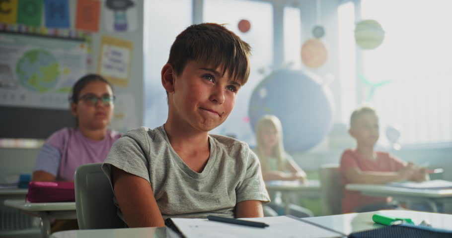 Primary School Boy Sitting at Desk, Raising Hand and Giving Correct Answer on Teacher's Question During Geography Lesson. Group of Young Boys and Girls Learning Science in Modern Colorful Classroom.
