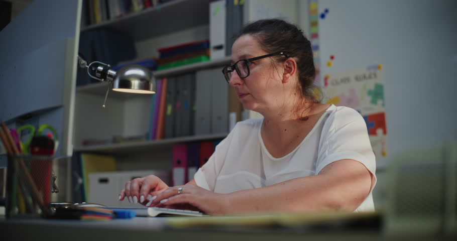 Elementary School Teacher Working on Computer, Checking Homework and Online Assignments During Distance Learning, Preparing Lesson Plan for Students. Woman Working at PC in Empty Classroom in Evening.