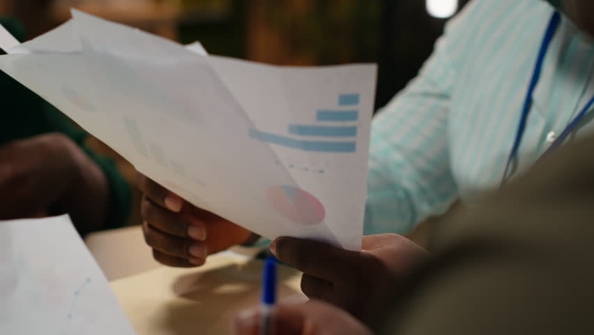Close up of diverse staff collaborating on data forecasting and analysis, exchanging documents late in the office. Updating stakeholders with predictions and other business intel. Camera B.