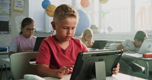 Elementary School Boy Sitting at Desk, Using Digital Tablet in Classroom. Team of Smart Diverse Kids Working on Laptops and Tablet Computers, Studying Computer Science, Basic Programming and Coding. - Powered by Shutterstock - Get 15% off with code: PIKWIZARD15