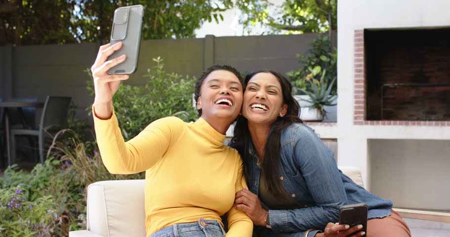 Diverse female friends lifting smartphone for selfie on garden patio, scrolling gallery. Outdoor, friendship, leisure, casual, lifestyle, vibrant, joyful