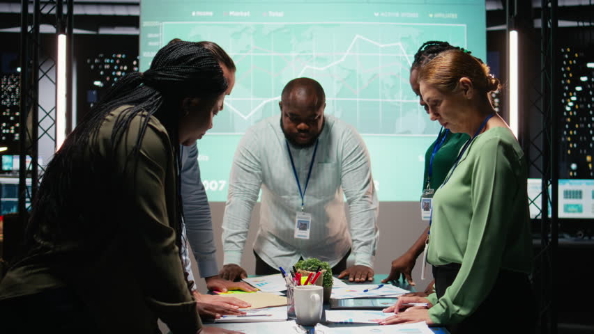Multiethnic team exchanging files and review forecasting diagrams at night, meeting in a quiet office space. After hours collaboration to prepare key project updates for shareholders. Camera A.