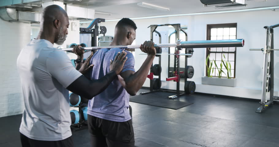 African American male trainer guiding trainee barbell squat rack spotting descent and ascent safely. Fitness, coaching, strength, stability, training, motivation, durability
