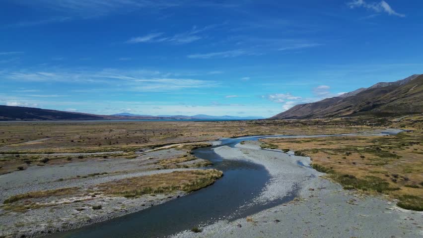 Drone footage following a braided river on a sunny day with mountains in the background, in South Island, New Zealand.