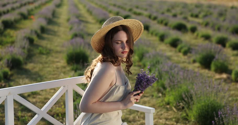 Beautiful young woman smelling lavender flowers on bench in field