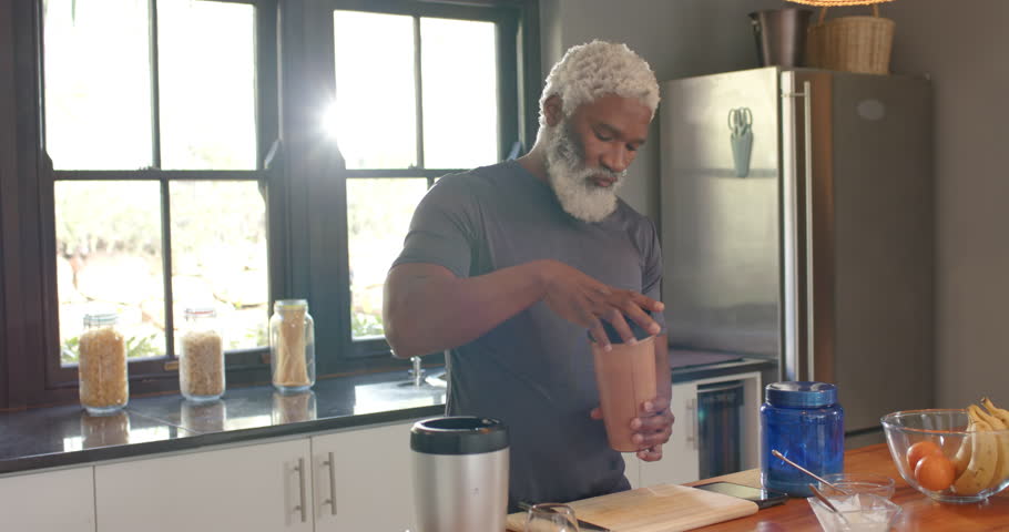 Waking senior African American man scooping supplement into blender cup mixing shake at counter. Healthy, nutrition, kitchen, lifestyle, wellness, modern, natural