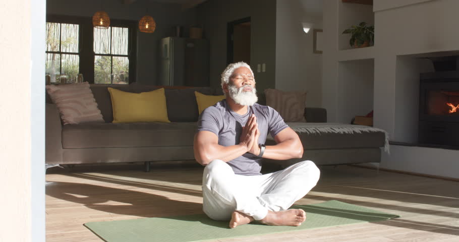 Sunlight guiding African American senior man sitting on green yoga mat meditating in living room. Serenity, mindfulness, wellness, relaxation, harmony, natural light, modern