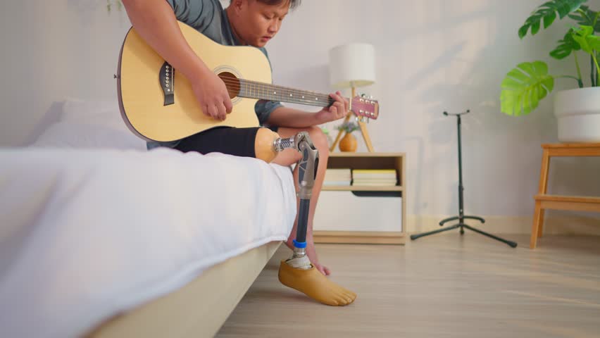 Asian man with prosthetic leg playing guitar on bed in bedroom at home. Attractive young male amputee wearing leg brace enjoying music and spend leisure relaxing time during recovery in cozy house.