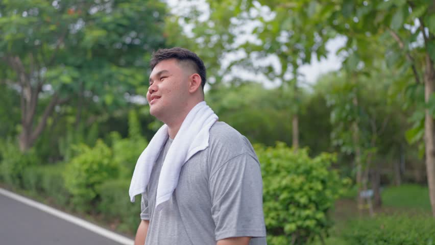 Asian plus-size man stretching body and exercise outdoors in garden. Attractive young overweight guy spend leisure time on weekend doing running workout to improve health and lose weight in the park.
