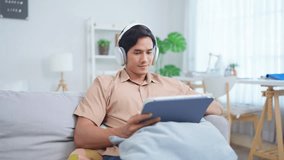 Asian young man using digital tablet and listen to music on headphones. Attractive guy sitting on sofa, using device working and browsing with happiness during holiday in cozy living room at home. - Powered by Shutterstock - Get 15% off with code: PIKWIZARD15