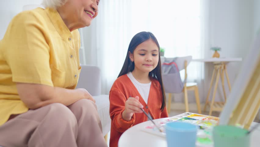 Asian loving grandmother support granddaughter with drawing at home. Caring elderly senior grandma encourage little girl's creativity, enjoying fun and educational art activity together in living room
