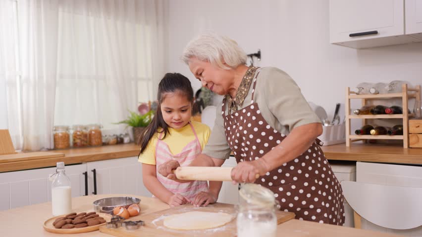 Asian granddaughter baking bakery with grandmother in kitchen at home. Happy family, adorable kid girl enjoys spending quality leisure time with caring senior mature grandma in cozy house together.