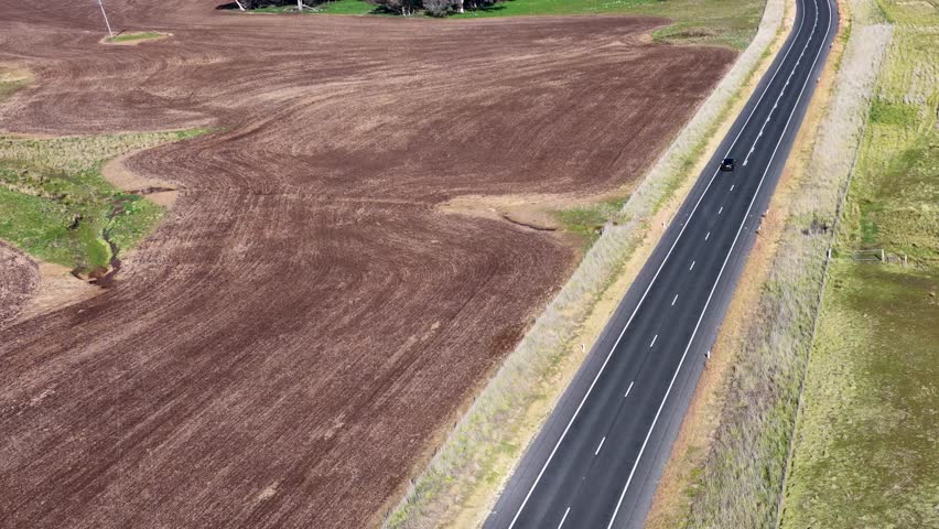 A white semi-truck travels along a winding rural highway bordered by plowed fields and sparse trees, captured in bright daylight from an aerial perspective