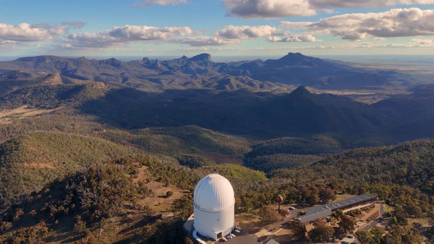 Aerial drone footage orbits a large white observatory dome atop a forested mountain ridge in Australia, with dramatic late afternoon sunlight and expansive views