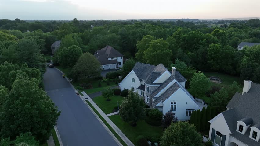 Upscale American suburb area. Large two-story homes with pitched roofs, stone and siding facades , surrounded by trees and winding roads in peaceful residential neighborhood. Aerial view at dusk.