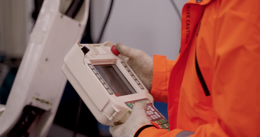 Close up of handheld robot controller with visible buttons and screen operated by gloved engineer during industrial robotics training session in automation lab for interface practice and diagnostics
