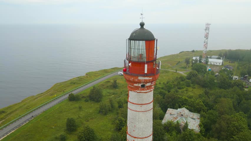 Aerial drone footage close up of a red colored historic lighthouse at a cliff seaside shoreline with a highway underneath it during a misty and foggy day In Pakri Paldiski at summer time. Gorgeous.