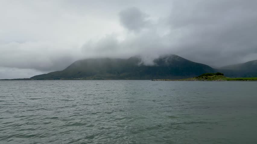 A scenic view of a tranquil fjord in Scandinavia with a small, rocky island and a large, misty mountain in the background under a cloudy sky.