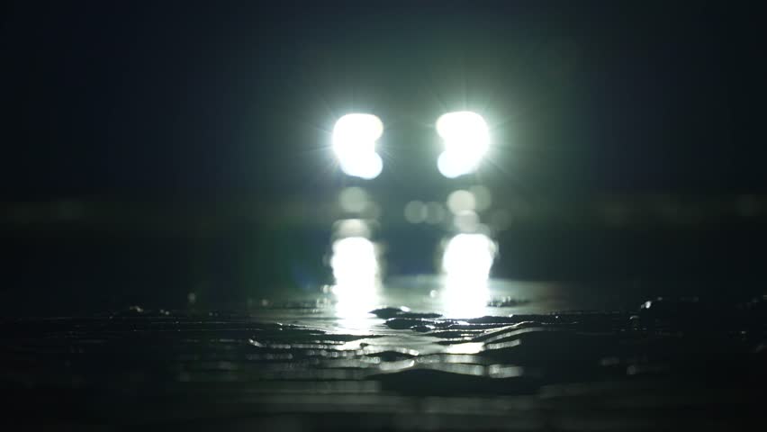 Mysterious car headlights shine through rainy darkness on empty road in cinematic horror atmosphere