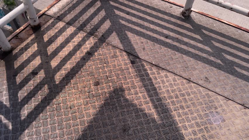 Close-up view of a textured metal floor with diamond plate pattern, illuminated by sunlight casting strong diagonal shadows from a railing.
