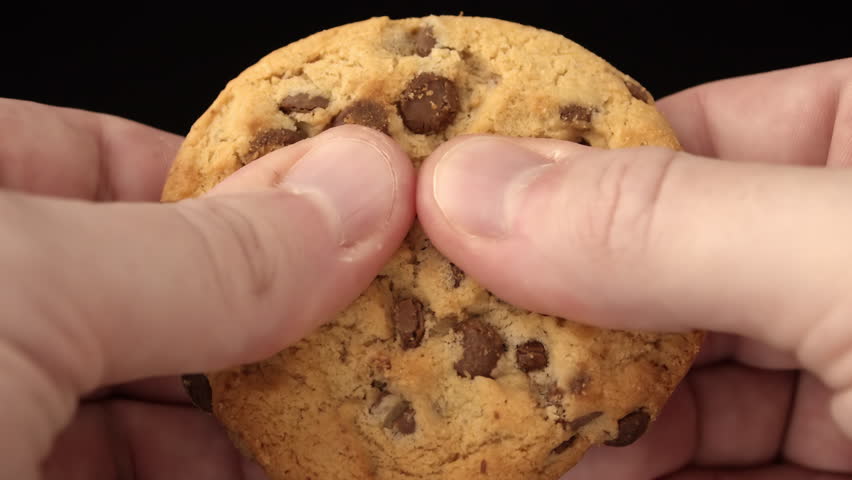 Chocolate chip cookie breaking in hands against black background with golden crumbs, captured as a snack break advertising moment