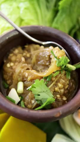 Hand uses a spoon to lift roasted green chili dip garnished with herbs from a rustic clay bowl, surrounded by fresh vegetables in natural daylight