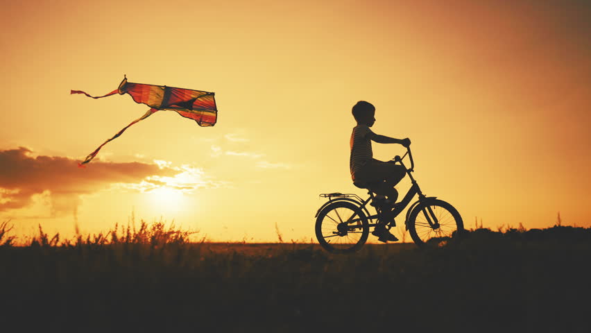 Flying kite moving behind little boy riding bike in summer weekend in nature. Cinematic slow motion shot on countryside with happy child, backlit silhouette against beautiful sky in sunset or sunrise