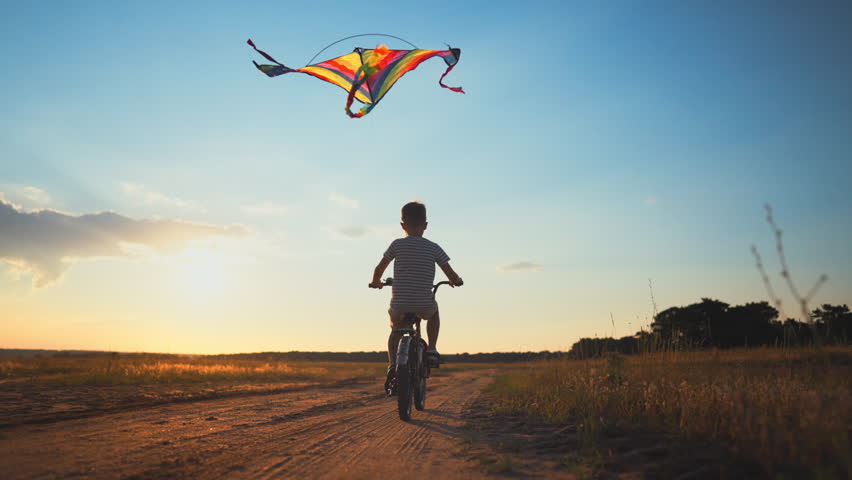 Child riding bicycle and flying kite in summer weekend , follow shot in nature. Happy childhood and carefree vacation in countryside, silhouette of little boy on bicycle in sunset or sunrise, freedom