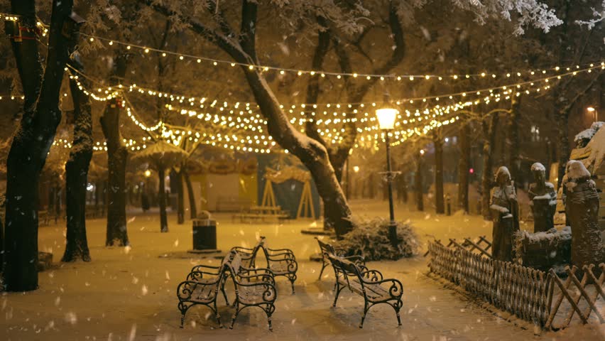 Snow falls in city decorated for Christmas and New Year holidays. Winter wonderland with twinkling lights and snow-covered trees in park at night