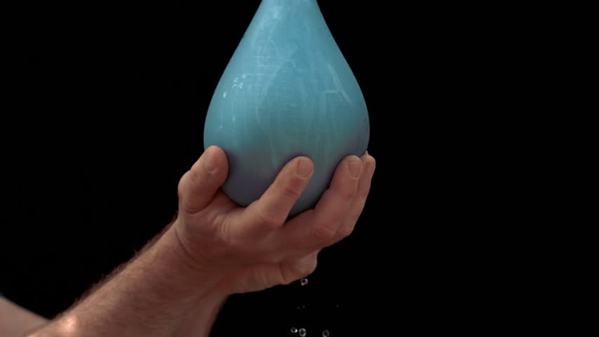 A hand forcefully pops a blue water balloon in slow motion, sending water flying against a dramatic black backdrop