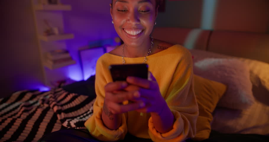 Young Cheerful Black Woman Sitting on a Bed at Home, Typing a Message for Social Network Audience on Her Smartphone. Neon Room Lighting Adds a Modern, Youthful Vibe. Zoom Out Footage