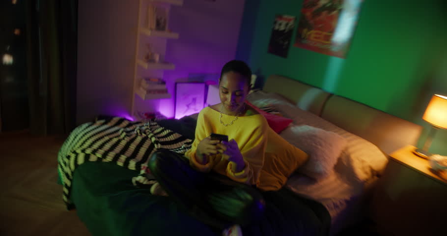 Young Female Sitting on a Bed in Her Gamer Room, Scrolling Through a Video Streaming App, Selecting Influential Content to Watch on Her Smartphone. Rotating Cinematic Robot Camera Movement