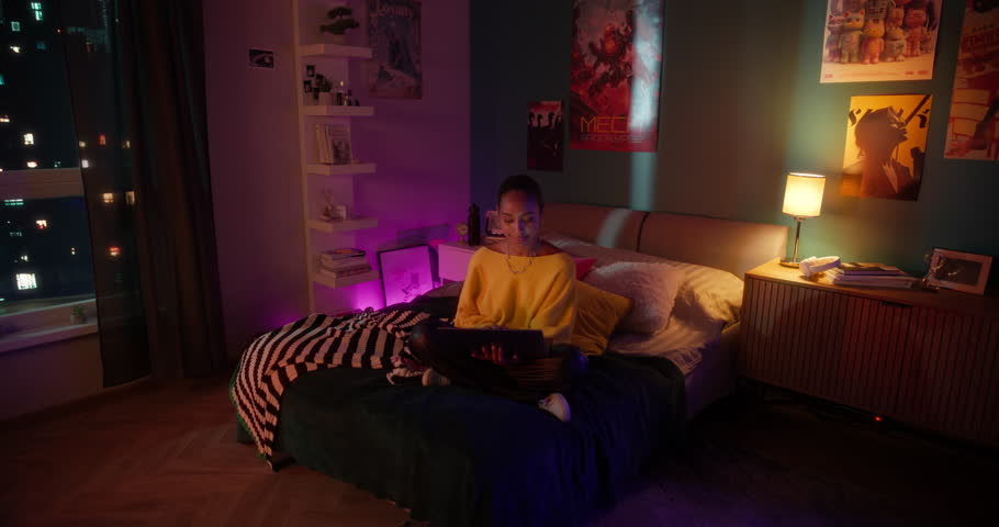 Young Black Female Sitting Cross-Legged on a Bed at Home, Working on Laptop Computer. Freelance Designer Doing Remote Work or Study for Exams. Swirling Robotic Camera Movement