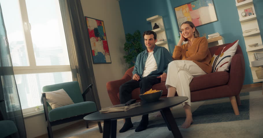 Two People Relaxing on a Couch, Watching TV and Chatting Between Scenes. Excited Daughter Running to the Young Couple, Sitting Down and Grabbing Popcorn. Casual Family Home Atmosphere