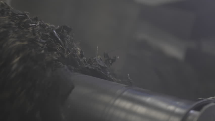 Massive rotating shaft slowly pushes shredded rubber into deep metal container. Scene shows intense steady industrial recycling process