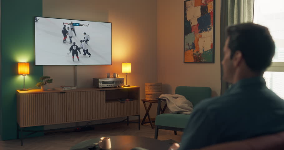 Young Man Watching Sports Live Stream with an Ice Hockey Match Broadcast. Two Teams Competing for the Championship Trophy. He is Sitting on a Couch at Home and Enjoying the Program