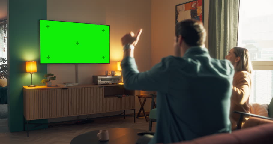 Young Couple Sitting on a Sofa and Watching a Sports Broadcast Channel TV with Green Screen Mock Up Template on Display. Man and Woman Cheering for Their Team and Athletes. Zoom Out Speed Ramp Footage