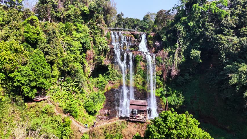 Aerial drone orbit reveals tiered Tad Gneuang Waterfall surrounded by lush jungle in Champasak Province, Laos, with misty cascades flowing beside a traditional wooden pavilion built on the cliffside.