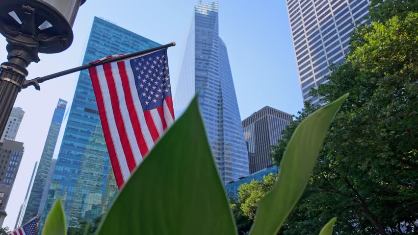 Low angle shot of American flag, modern glass skyscrapers, green foliage in foreground, symbol of patriotism, finance, and business in urban setting, New York City, USA