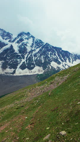 Majestic mountain scenery showing a steep green hillside leading up to rugged, snow-streaked peaks under a cloudy sky. Vertical handheld footage as seen from ropeway